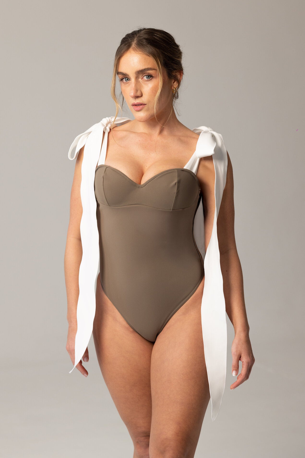 Woman wearing a taupe one-piece swimsuit with large white shoulder ties, elegant and modern design, featuring the luxury swimwear piece MATHEA.