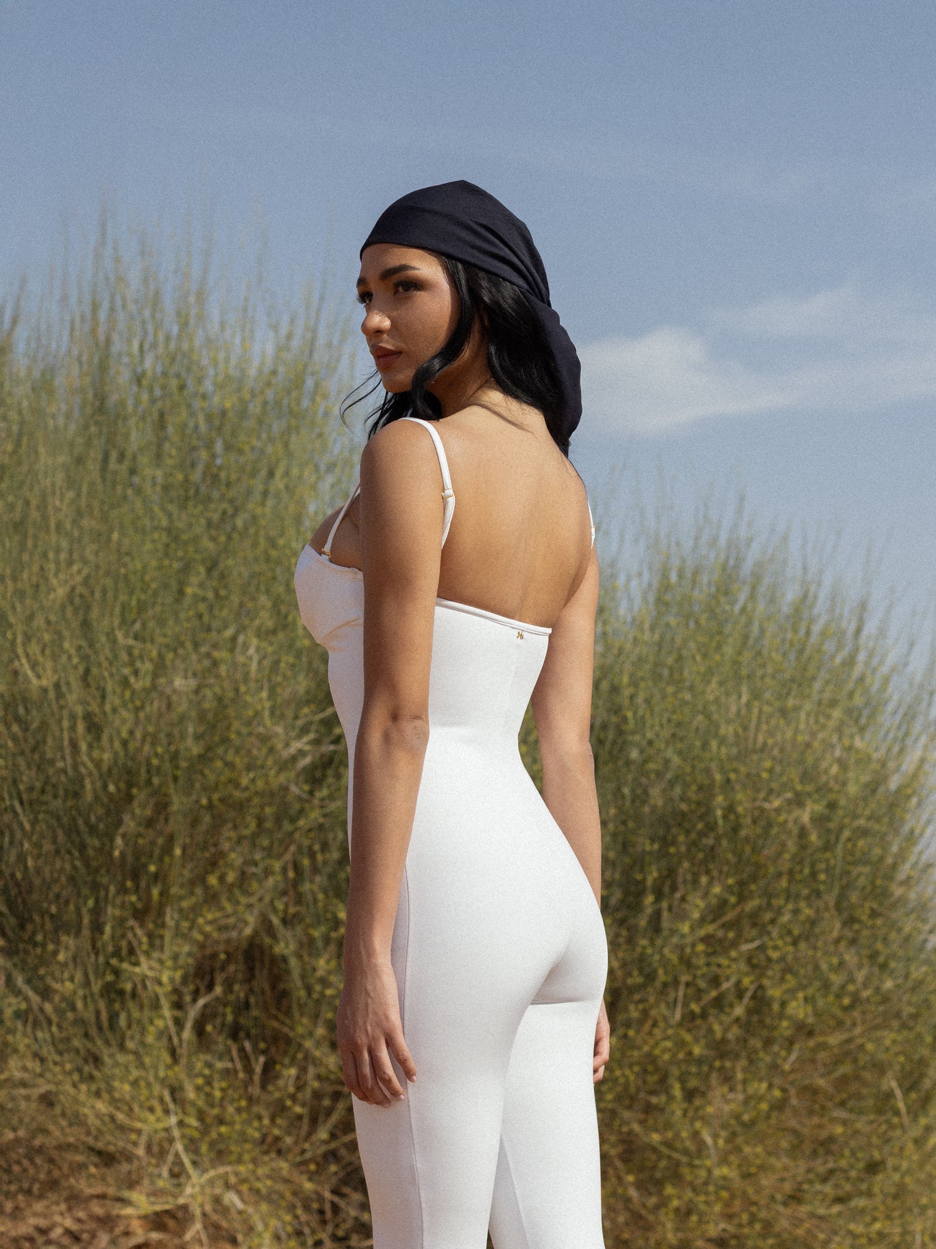 Woman wearing a sleek white fitted outfit and a black headscarf standing outdoors with greenery and blue sky in the background, showcasing NURA BODY luxury swimwear.