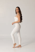 Woman wearing a sleek white one-piece swimsuit standing barefoot against a plain background, showcasing the elegant and minimalist design of the NURA BODY luxury swimwear.