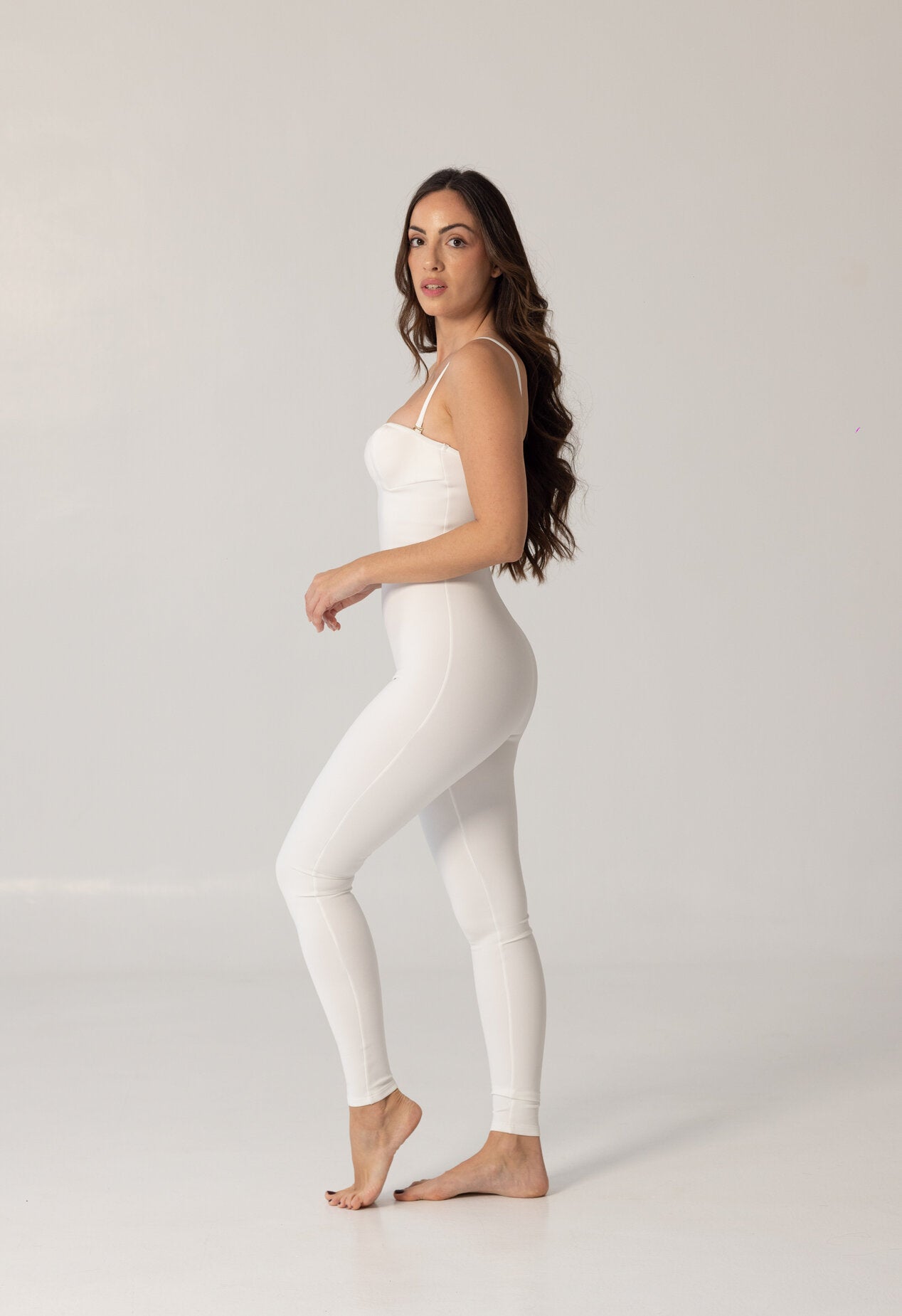 Woman wearing a sleek white one-piece swimsuit standing barefoot against a plain background, showcasing the elegant and minimalist design of the NURA BODY luxury swimwear.