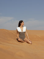 Woman seated on sand dunes wearing ARIEL luxury swimwear with a deep V neck and contrasting white and beige colors under a clear blue sky.