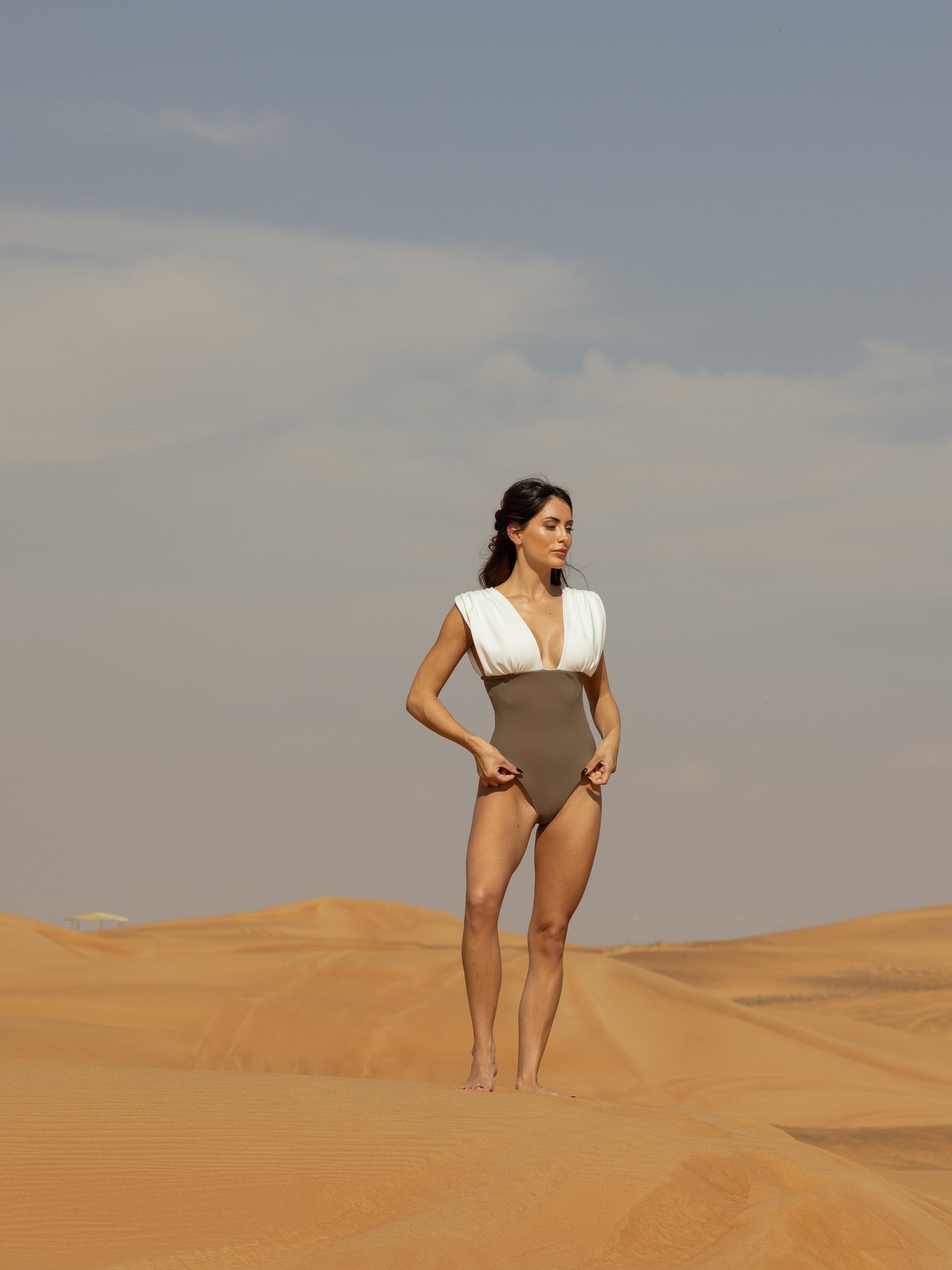 Woman standing in a desert wearing a stylish ARIEL one-piece swimsuit with a deep V-neck and structured white shoulder detail under a clear sky