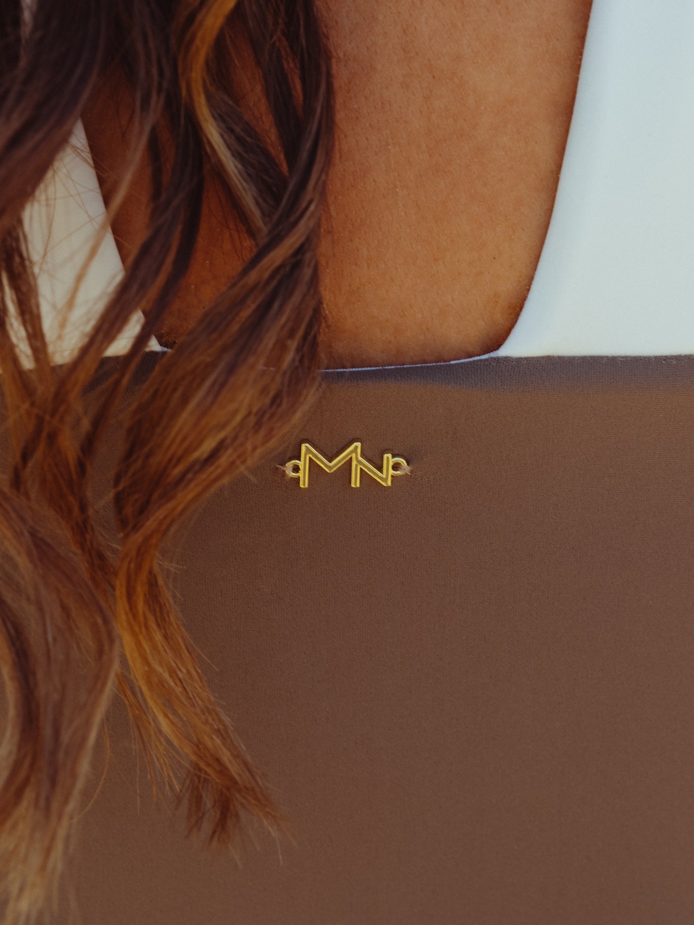 Close-up of a brown luxury swimwear piece with golden Maison Nura logo detail on the back, worn with visible white strap, highlighting the ARIEL collection.