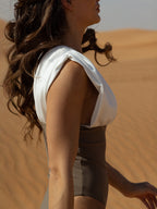 Close-up side view of a woman wearing the ARIEL luxury swimwear one-piece swimsuit in a desert setting, featuring a white draped shoulder design and brown fitted body.