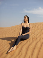 Woman wearing a sleek black one-piece swimsuit from NURA BODY, sitting barefoot on sand dunes under a clear sky, embodying elegance and luxury swimwear style.