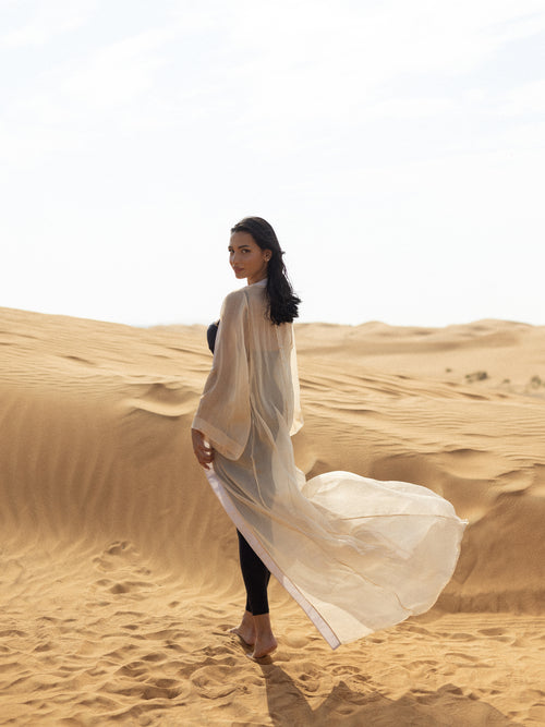 Woman wearing flowing light beige beach cover-up standing on sandy desert dunes, showcasing the elegant and modest luxury swimwear piece ISAURA in natural sunlight.