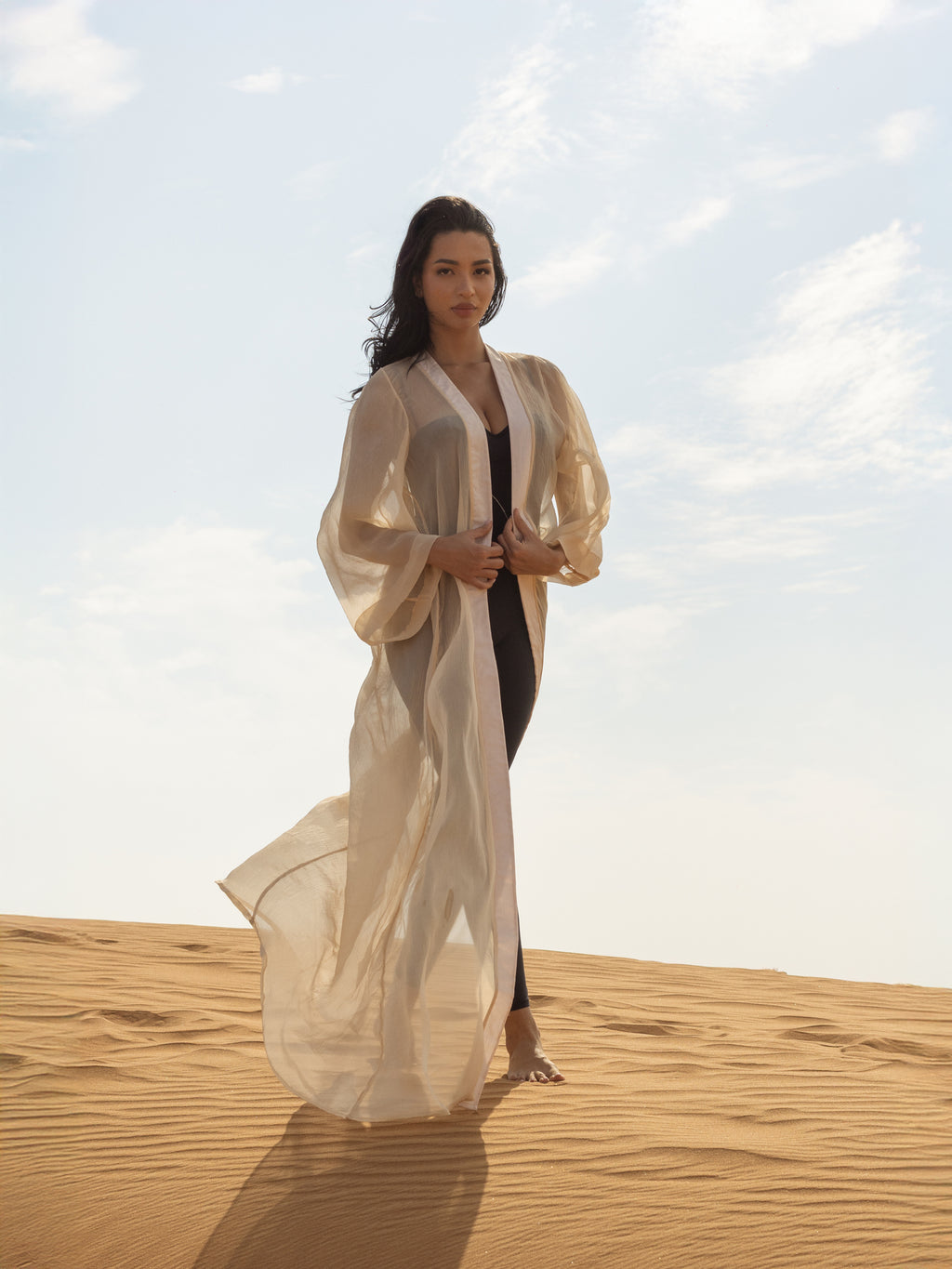Woman wearing a flowing sheer beige cover-up called ISAURA over a black outfit, standing barefoot on sand dunes under a bright sky.