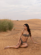 Woman wearing BRUNA luxury swimwear in muted earth tone sitting on sandy desert landscape with sparse grass under cloudy sky creating serene and elegant mood.