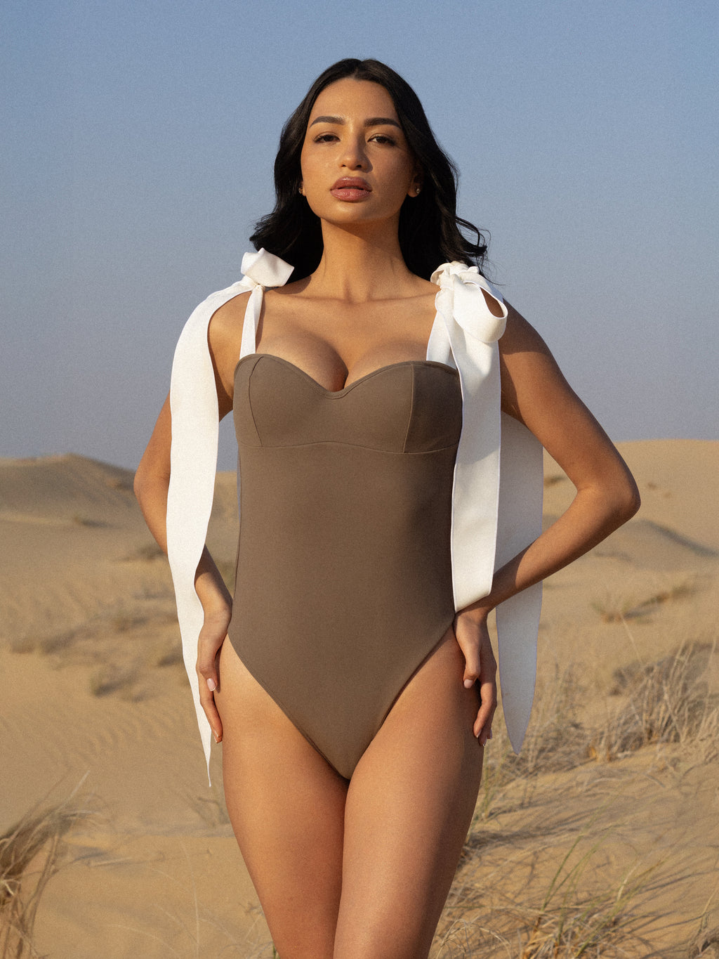 Woman standing in desert wearing a taupe MATHEA one-piece swimsuit with white ribbon shoulder ties, showcasing luxury swimwear style against a sandy dune backdrop.