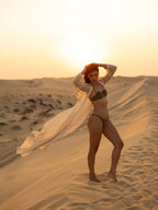 Woman wearing JANNAH luxury swimwear standing on sand dunes at sunset with a flowing cover-up cape, showcasing premium resort wear in a serene desert setting.