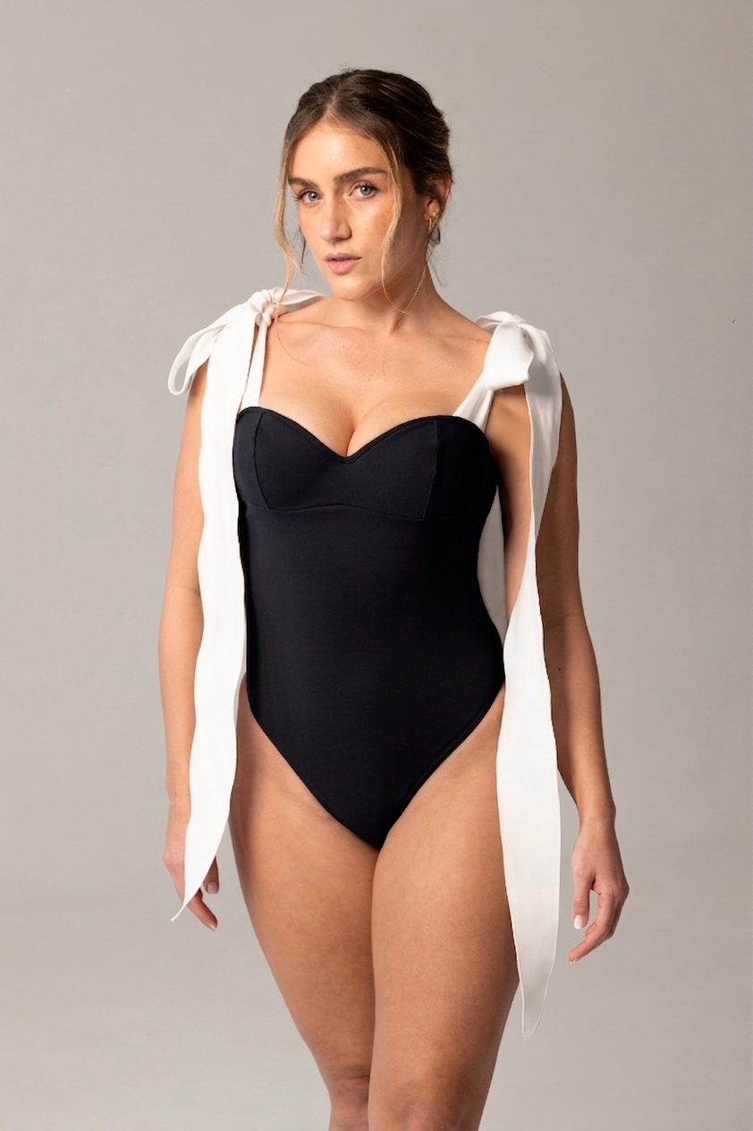 Woman wearing a black MATHEA one-piece swimsuit with white shoulder ties, showcasing a luxury swimwear design perfect for elegant resort wear.